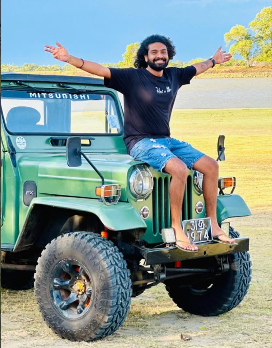 Mr. Maleesha, founder of Maleesha Lanka, sitting on the bonnet of a green Mitsubishi safari jeep with his arms spread wide, beside a lake in the dry zone