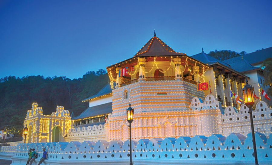The Temple of the Tooth in Kandy lit with strings of lights at dusk, with forested hills behind