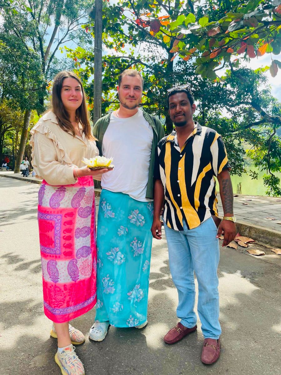 Two guests wearing sarongs, holding a lotus offering, with their driver-guide beside Kandy lake