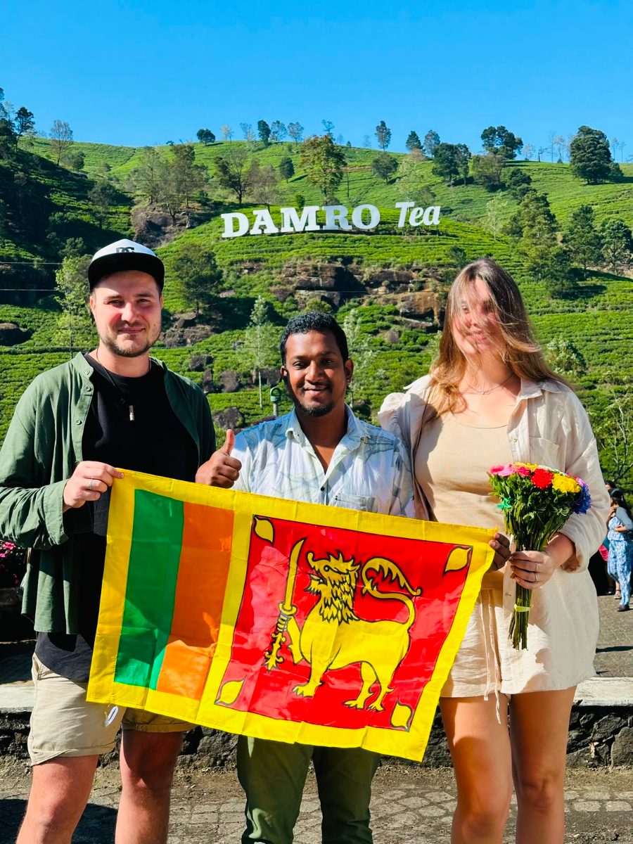 Two guests holding a Sri Lankan flag with their driver-guide in front of the tea slopes at a hill-country estate